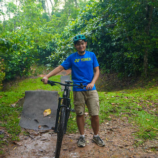 Person with a bicycle on a forest trail wearing a blue shirt and helmet.